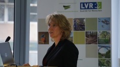 Dr. Martina Gelhar begrüßt die Teilnehmenden zum 14. Forum Kulturlandschaft und Naturschutz. Sie steht am Rednerpult. Im Hintergrund ist das Banner des LVR-Netzwerks Kulturlandschaft mit den Biologischen Stationen im Rheinland zu erkennen.