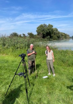 Zwei Menschen auf einer Wiese vor einem See, im Vordergrund eine Videokamera, die Frau hält ein Mikrofon