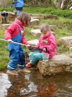 Zwei Mädchen am Wasser. Sie tragen wetterfeste Kleidug und Gummistiefel. Sie haben beide einen Kescher in der Hand und versuchen Wassertiere einzufangen.